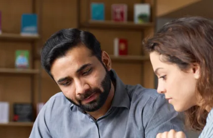 Man and woman sitting at table looking together at a notepad while he holds a sheet of paper and she is holding a pencil and writing. Both are dressed professionally with him in a collared shirt and her in a striped blouse. He has a stylish haircut and she wears her hair down and blown-out.