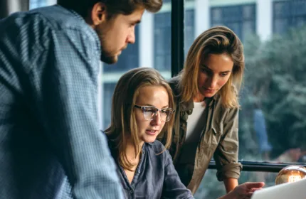 Three colleagues gathered around a laptop, collaborating on a project in a modern office with natural light.
