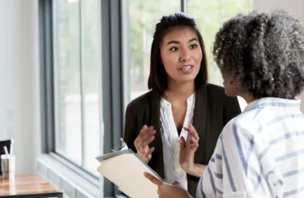 Two coworkers stand in a bright office near large windows, reviewing printed documents together during a discussion.