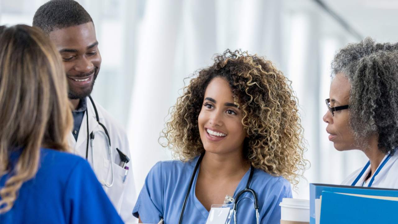 A group of medical professionals in scrubs and lab coats standing in a bright hospital corridor, engaged in conversation. One person holds a tablet, another carries a clipboard, and a third has a coffee cup, symbolizing collaboration and teamwork in healthcare settings.