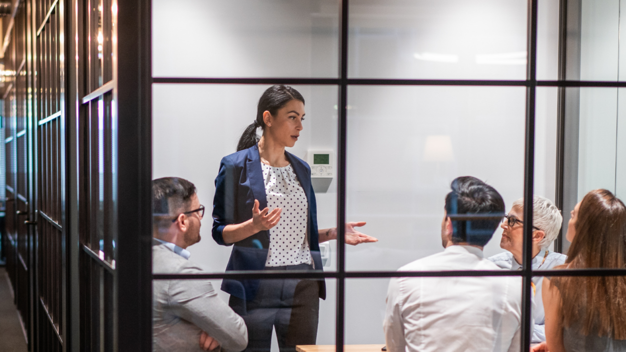 A modern glass-walled conference room with several professionals seated around a wooden table while another person stands and speaks, gesturing during a presentation. Laptops and documents are visible on the table, representing collaboration and strategy planning for sales and business development.