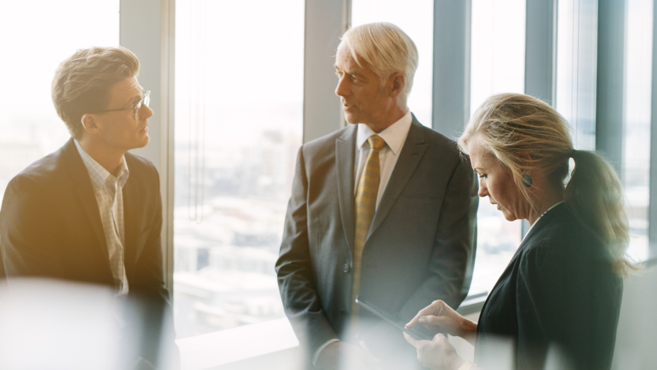 Three professionals standing near large floor-to-ceiling windows in a modern office, engaged in a discussion.