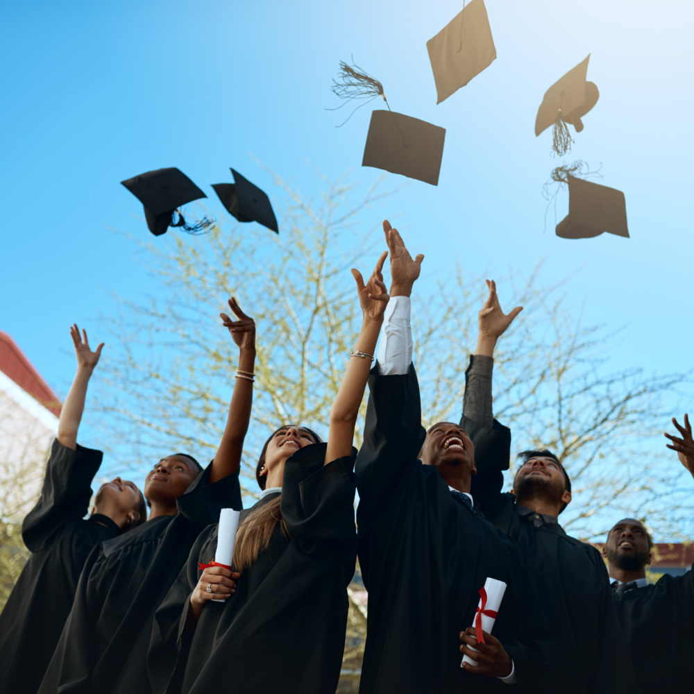 Graduates wearing black caps and gowns toss their mortarboards into the air outdoors, holding diplomas in hand. The scene represents academic achievement and celebration in an educational setting.