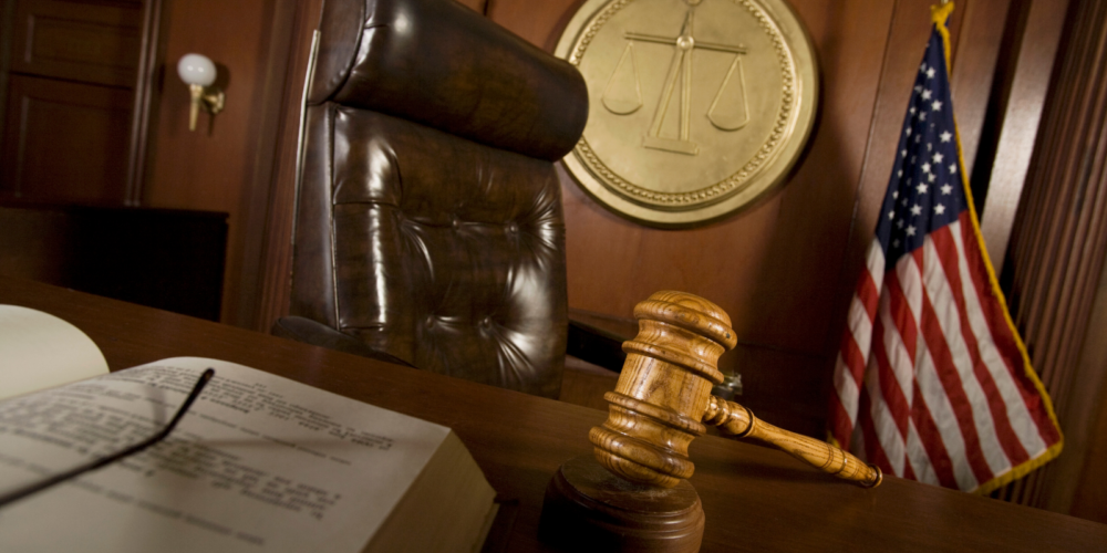 A courtroom scene features a legal professional presenting documents to a judge behind a wooden bench, with law books and traditional furniture, symbolizing litigation.