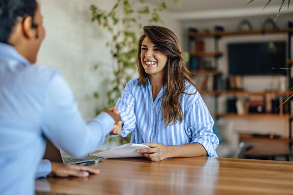 Two professionals shake hands across a desk with paperwork, representing a successful interview, hiring decision, or business partnership.